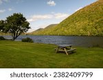 An empty campsite next to a river with a lush yellow and green colored hillside of trees. The sky is blue with clouds. There