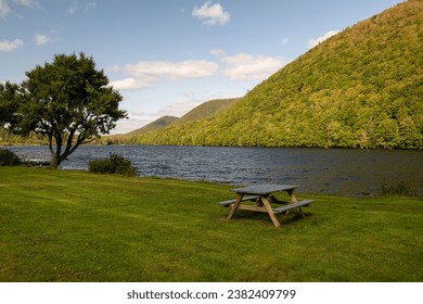 An empty campsite next to a river with a lush yellow and green colored hillside of trees. The sky is blue with clouds. There's a picnic table on the grass-covered park campground with no people. 