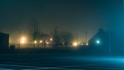 An Empty Car Park With Street Lights Glowing In The Distance On A Mysterious Moody,  Foggy Atmospheric Winters Night
