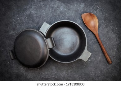 Empty Cast Iron Pot With Lid And Wooden Spoon On Black Isolated Background Top View. Empty Iron Pot On Grunge Black Table, Overhead View.