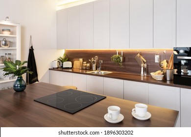 Empty ceramic glass stove on wooden countertop table against modern interior with white kitchen cupboards, sink, electric oven and kitchenware supplies
