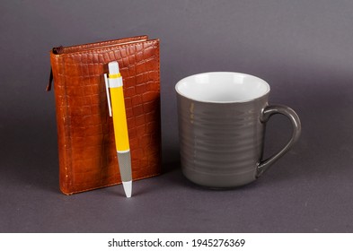 Empty ceramic mug, pen and notebook on gray background. New dark gray mug, yellow pen and brown notebook. Selective focus.