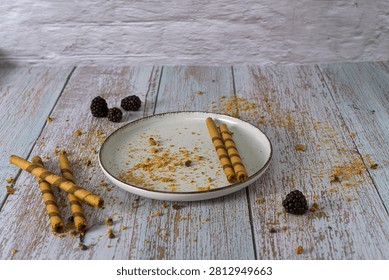 Empty ceramic plate with leftover cookie crumbs, rolled wafers, and fresh blackberries on a rustic wooden table after eating a delicious dessert