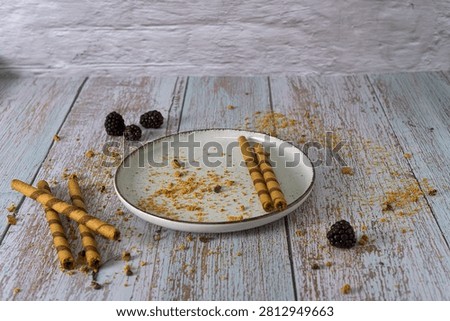 Empty ceramic plate with leftover cookie crumbs, rolled wafers, and fresh blackberries on a rustic wooden table after eating a delicious dessert
