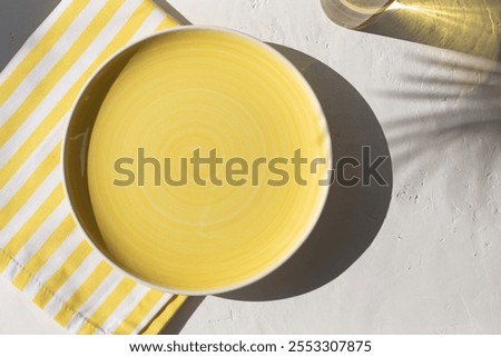 Empty ceramic yellow plate on the white plaster background with yellow striped pattern napkin or towel with tropical palm leaves shadows. Summer table setting. Copy space