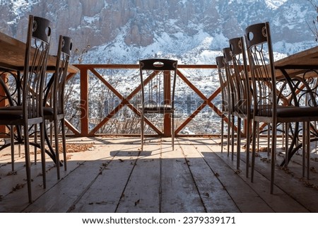 Empty chairs at tables on left and right and one chair in the middle put on a sunlit wooden terrace overlooking snowymountains