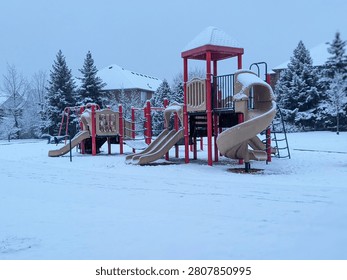 An empty children's playground structure with red poles and tan slides sits covered in fresh winter snow. Snow-coated evergreen trees and suburban houses stand in the background under a grey sky.