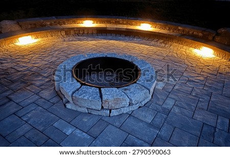 Empty circular stone fire pit on a paved backyard patio at night with warm landscape lights, curved seating wall and modern hardscape design.