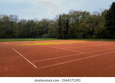 An empty clay tennis court sits peacefully in the woods, surrounded by vibrant autumn foliage, creating a serene atmosphere for leisurely games and quiet reflection. - Powered by Shutterstock