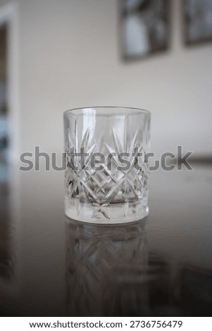 An empty clear crystal drinking glass featuring intricate cut patterns like starbursts and diamonds rests on a dark, reflective table surface. Bright room light illuminates its elegant details