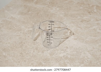 An empty, clear plastic measuring cup sits on a beige marble kitchen counter. The jug has markings for milliliters, liters, sugar, flour. Minimalist composition with soft lighting neutral colors.