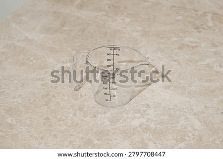 An empty, clear plastic measuring cup sits on a beige marble kitchen counter. The jug has markings for milliliters, liters, sugar, flour. Minimalist composition with soft lighting neutral colors.