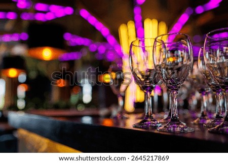 Empty clear wineglasses stand in rows on bar counter at nightclub, blurred purple neon and yellow lights on background. Clean cocktail glasses ready for celebrate party.