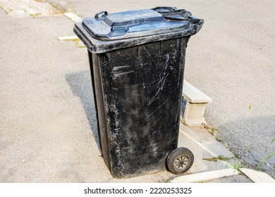 Empty And Closed Dirty Plastic Black Trash Bin With Wheels On A Sidewalk