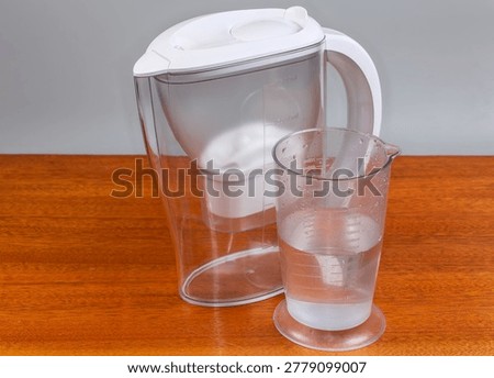 Empty closed plastic transparent tap water purifier jug and plastic measuring cup with water on a wooden table on a gray background