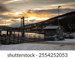 Empty closed port of a traject to the small islands. Mountain with trees and houses in the background. Snow on the land. Cloudy sky with sunshine in the sunrisesunset. Stornes, Harstad, Norway.