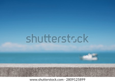 An empty concrete surface with a blurred image of a boat in the distance on a calm sea against a bright sky. A banner for showcasing summer products. Concept of vacation and serenity. Front view, copy space.