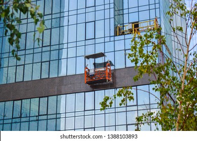 Empty construction suspended cradle / trolley hanging at the wall of a modern steel and glass building. Scaffold elevator at the wall of a scyscraper with few tree brnches on the foreground. 