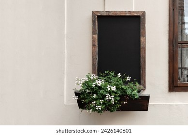 Empty dark wooden chalkboard mockup, flowers in outdoor planter, hanging on wall facade of authentic retro cafe. Menu template for old cafe or restaurant.