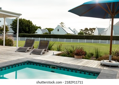 Empty Deckchairs And Umbrella At Poolside And Houses With Solar Panels On Rooftop Against Sky. Copy Space, Nature, Building, Solar Energy, Electricity, Green Technology And Sustainability Concept.