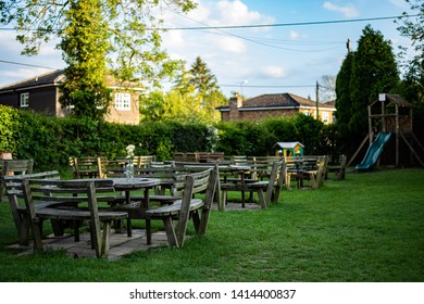 Empty English Pub Garden In The Early Evening. Lots Of Tables With Chairs And A Children's Play Area In The Background. Blue Skies With Small Clouds. Trees And Houses Are Present.