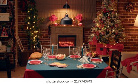 Empty festive christmas dinner table inside decorated living room with holiday garlands and dinnerware. Interior of traditional and authentic season cozy setting celebrating religious event.