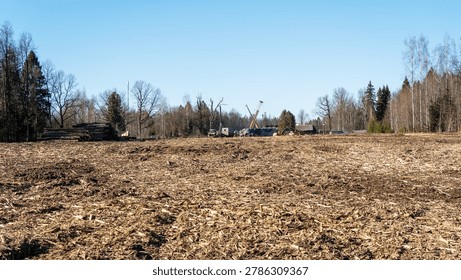 Empty field under clear blue sky. Brown stubble covers earth after harvest. Trees stand bare against distant horizon. Crane and trucks suggest recent construction