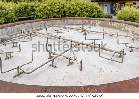 An empty fountain with a complex pipe system can be seen in the center of a well-kept park. The rusted pipes and chipped plaster at the edge of the pool indicate disuse or the need for renovation.