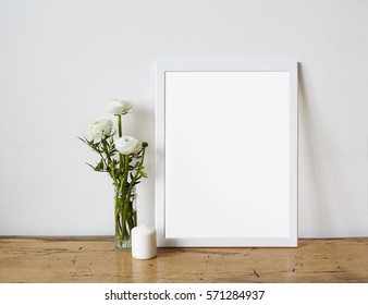 Empty frame mockup for design presentation, bouquet of flowers ranunculus and white candle on a white wall background and wooden table. Romantic minimalism design.