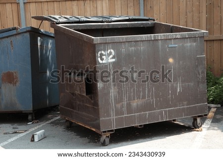 empty garbage dumpster with lid open and another dumpster behind it, surrounded by wood fence in parking lot, full dumpster in shot