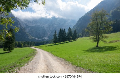 Empty Gravel Hiking Trail Leads Across A Lush Green Meadow And Towards A Majestic Mountain In Jezersko, Slovenia. Scenic Shot Of A Path Running Across Empty Pastures Underneath A Rocky Ridge In Europe