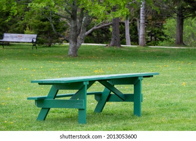 An Empty Green Wooden Picnic Table That's Wheelchair Accessible On One Side For A Disabled Or Handicapped Chair. There's A Park Bench In The Background With Large Trees And Green Grass.