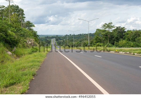 Empty highway downhill with street lamp, trees\
and white clouds.