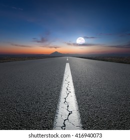 Empty Highway Leading To The Mountains Through The Desert Against The Rising Full Moon At Night.