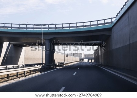 Empty highway road passing under modern concrete overpass and bridge structure. Urban transport infrastructure with multilane asphalt roadway, barriers and flyover viaduct in daylight travel concept.