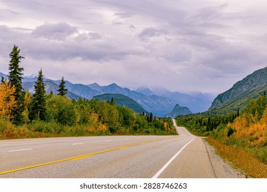 An empty highway winds through fall-colored forest toward distant peaks along the Glenn Highway, Alaska.
