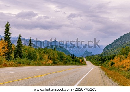 An empty highway winds through fall-colored forest toward distant peaks along the Glenn Highway, Alaska.
