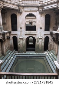 An empty historic stepwell interior features symmetrical stone arches, carved details, and a still water pool at the center, monument in Ahmedabad