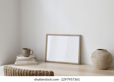 Empty horizontal picture frame mockup in minimalist interior. Wooden table, blurred rattan chair. Vase with cup of coffee. White wall background. Landscape template of artwork, painting, poster. - Powered by Shutterstock