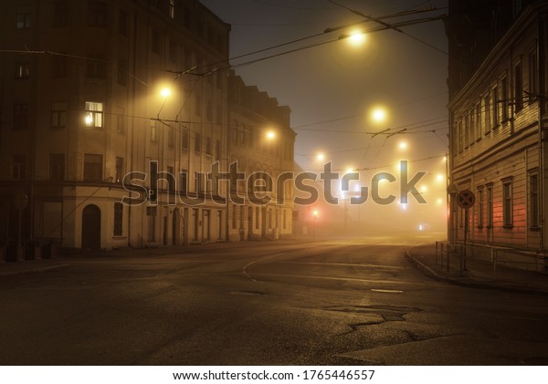 An empty illuminated asphalt\
road through the old historical buildings and houses in a fog at\
night. Street lights (lanterns) close-up. Riga, Latvia. Dark\
cityscape