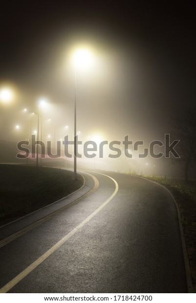 An empty illuminated\
bicycle road with a sharp turn in a fog at night. Lanterns\
close-up. Bridge in the background. Recreation and healthy\
lifestyle theme. Riga,\
Latvia