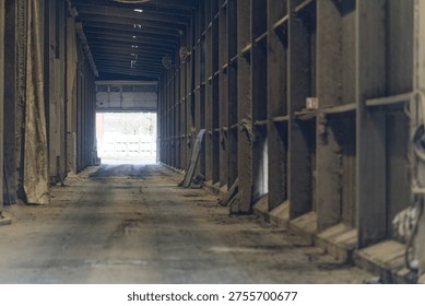 An empty industrial warehouse corridor featuring metal beams and a concrete floor leading to an open exit door with natural light outside.