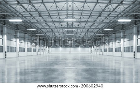 Empty interior space inside a modern industrial building, factory, or large warehouse, designed for manufacturing and production operations. The structure features a steel frame truss system with metal sheet walls and roofing, creating a strong and durable industrial environment in a dark gray tone. The space includes a clean, polished concrete floor with a smooth finish, suitable for industrial product display, machinery setup, storage, or logistics operations. The open layout provides flexibility for manufacturing processes, warehouse organization, and distribution activities, making it ideal for various industrial and commercial applications. This minimal and spacious setting works well as a background for industry, engineering, and production concepts, highlighting themes such as efficiency, infrastructure, and modern factory design.