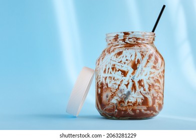 Empty Jar Of Chocolate Nut Spread With A Spoon On A Light Blue Background. Favorite Delicacy, Traditionally For Breakfast, Popular Sweet Paste For Sandwiches And Baked Goods.