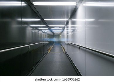 Empty jet bridge with blank-panel reflective walls, one side light one side dark. No-skid rubber floor and railings for safety. Secure area.