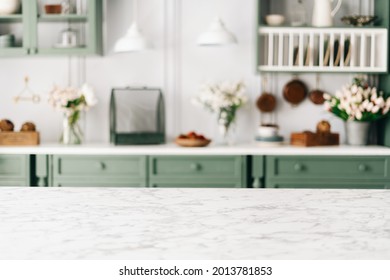 Empty Kitchen Island With Marble Surface In Foreground, Green Vintage Countertop With Drawers And Pendant Lights Hanging Above, Lots Of Flowers In Jars, Blurred Background