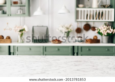 Empty kitchen island with marble surface in foreground, green vintage countertop with drawers and pendant lights hanging above, lots of flowers in jars, blurred background