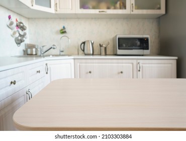 Empty Kitchen Island With Wooden Surface In Foreground, White Modern Countertop With Drawers And Microwave Oven, Lots Of Cups On Wall, Blurred Background