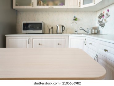 Empty Kitchen Island With Wooden Surface In Foreground, White Modern Countertop With Drawers And Microwave Oven, Lots Of Cups On Wall, Blurred Background