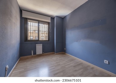 An empty living room with a blue wall, cheap wooden floors and oak doors and a bay window with steel bars